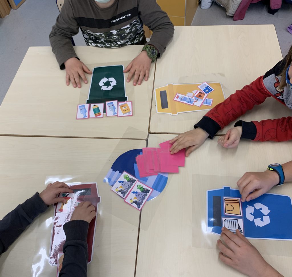 Niños jugando con cartas de reciclaje. Aparecen varios contenedores (verde, azul, amarillo y marrón) y una baraja de cartas boca abajo. Al dar la vuelta a la carta aparece la imagen de un objeto a reciclar