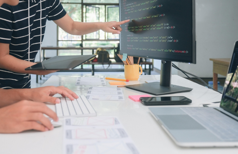 Dos personas colaboran frente a un monitor que muestra código de programación, rodeadas de bocetos de diseño y dispositivos electrónicos sobre una mesa de trabajo.